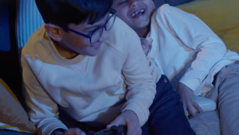 Two-Young-Boys-At-Home-Having-Fun-Playing-With-Computer-Games-Console-On-TV-Holding-Controllers-Late-At-Night